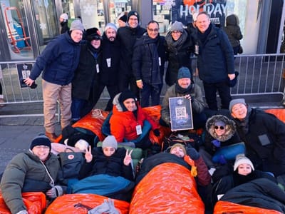 Group of about fifteen World's Big Sleep Out participants posing in Times Square, some standing in winter coats and others lying in orange sleeping bags on the sidewalk, one holding a framed event sign