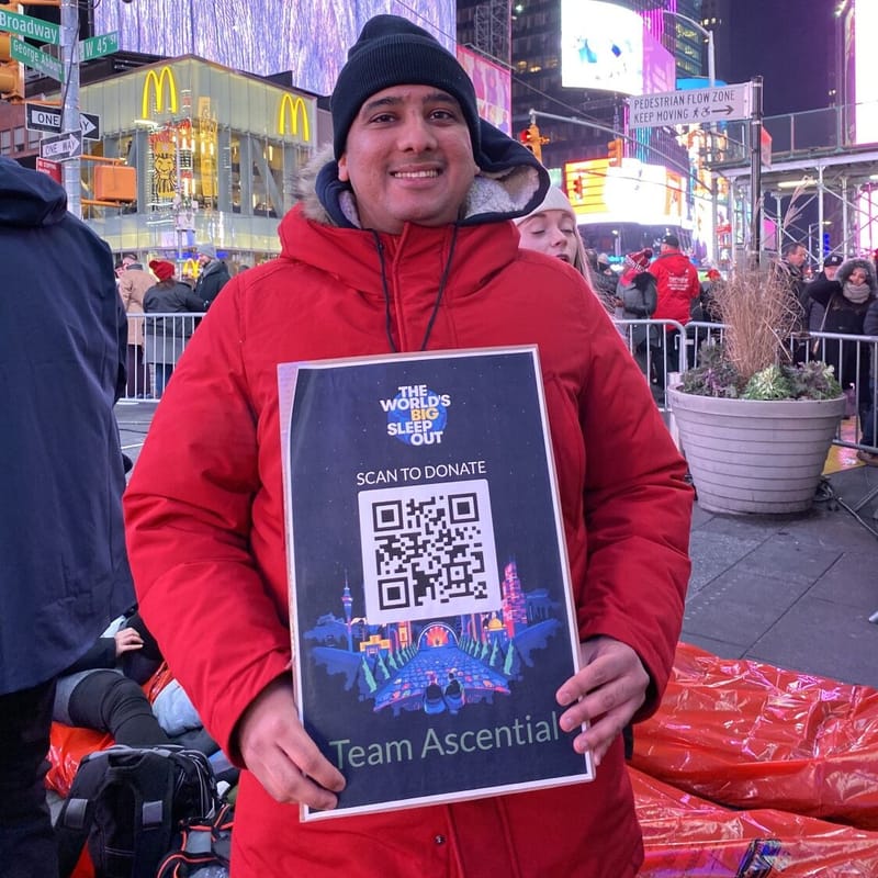 A World's Big Sleep Out participant in a red winter coat holds a Scan to Donate poster with a QR code in Times Square